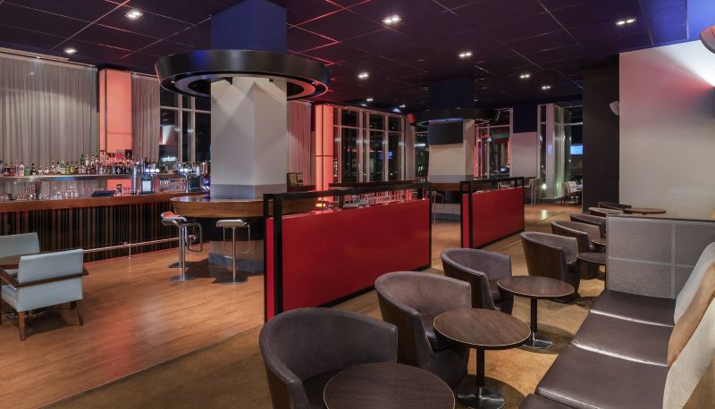 The inside of a bar, at the Novotel London ExCel. There are brown chairs and tables in front of a bar that serves drinks.
